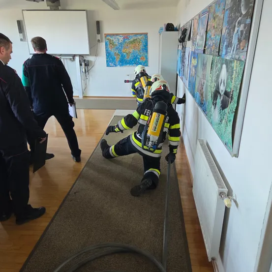 Mehrere Feuerwehrleute in Schutzkleidung kriechen durch einen Flur. Sie befinden sich neben einer Wand mit einer Tafel, Karten und einem Heizkörper. Drei Männer in Uniformen beobachten sie.