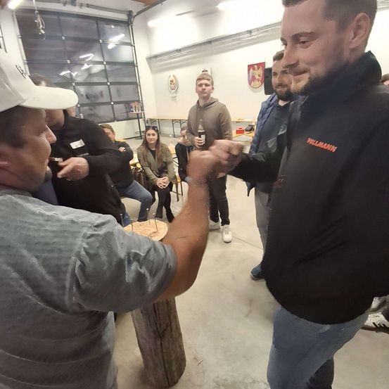 Zwei Männer treten in einem sozialen Umfeld in einem Armdrücken-Wettbewerb gegeneinander an, während andere zuschauen, einige sitzend und Getränke in der Hand. Der Mann auf der rechten Seite trägt ein schwarzes Shirt mit orangem Text.