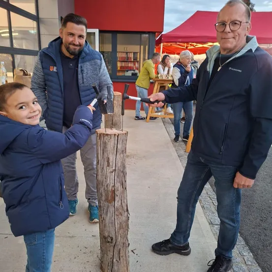 Zwei Männer und ein Kind verwenden Hämmer, um einen Baumstamm auf dem Bürgersteig zu spalten. Hinter ihnen stehen Menschen um einen Tisch mit Getränken. Ein rotes Zelt ist im Hintergrund zu sehen.