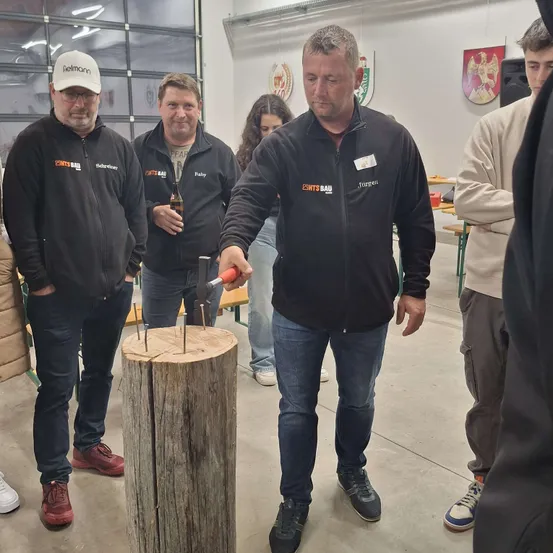 Ein Mann schlägt Nägel in einen Holzstumpf in einer Werkstatt, beobachtet von anderen. Er trägt einen schwarzen Mantel mit Namensschild. Zwei Männer in Brillen beobachten von der Seite.