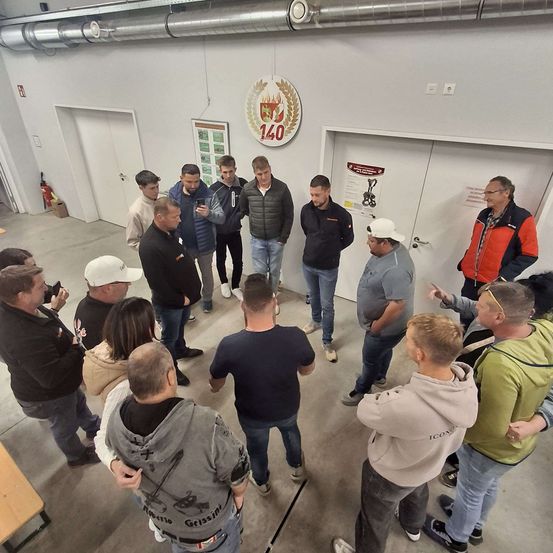 Eine Gruppe von Männern und Frauen steht in einem Kreis in einem Raum. Ein Mann hält ein Handy und einige Leute machen Fotos. Ein Feuerlöscher ist an der Wand montiert.