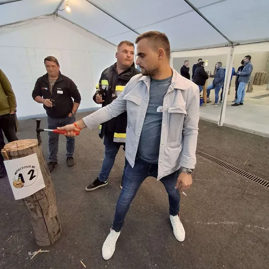 Ein Mann steht in einem Zelt und hält einen Hammer in der Hand, um einen Holzpfosten mit der Nummer 12 zu treffen. Hinter ihm beobachten zwei Männer. Andere sind im Hintergrund, einige stehen, andere sitzen.