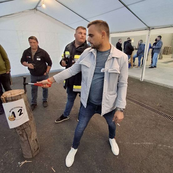 Ein Mann steht in einem Zelt und hält einen Hammer in der Hand, um einen Holzpfosten mit der Nummer 12 zu treffen. Hinter ihm beobachten zwei Männer. Andere sind im Hintergrund, einige stehen, andere sitzen.