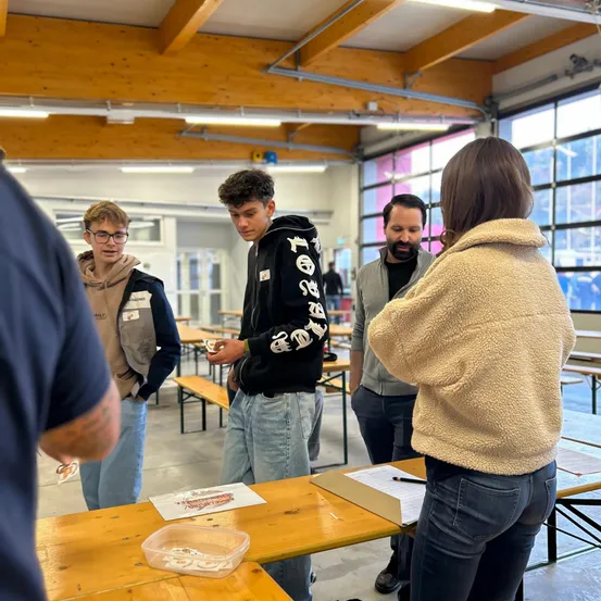 Eine Gruppe von Menschen steht um einen Tisch herum, einige unterhalten sich, während ein anderer ein Stück Papier in der Hand hält und einen Plastikbehälter mit Gegenständen betrachtet. Der Raum hat Holzbänke, Glasfenster und eine Holzdecke.