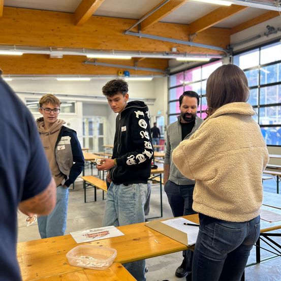 Eine Gruppe von Menschen steht um einen Tisch herum, einige unterhalten sich, während ein anderer ein Stück Papier in der Hand hält und einen Plastikbehälter mit Gegenständen betrachtet. Der Raum hat Holzbänke, Glasfenster und eine Holzdecke.