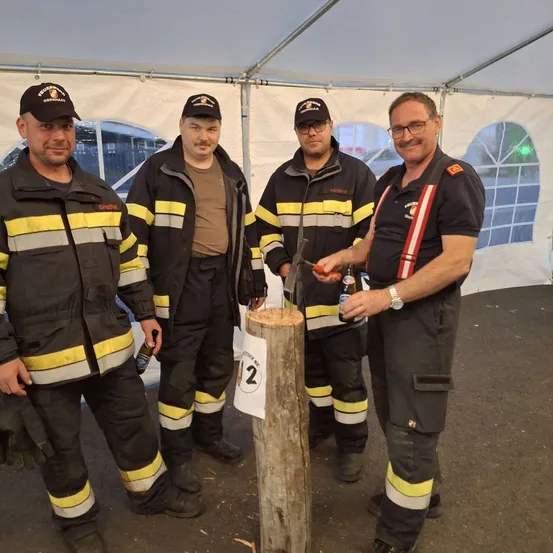 Vier Männer in Feuerwehruniformen stehen um einen Holzpfosten. Ein Mann hält eine Flasche und ein anderer einen Hammer. Sie lächeln.