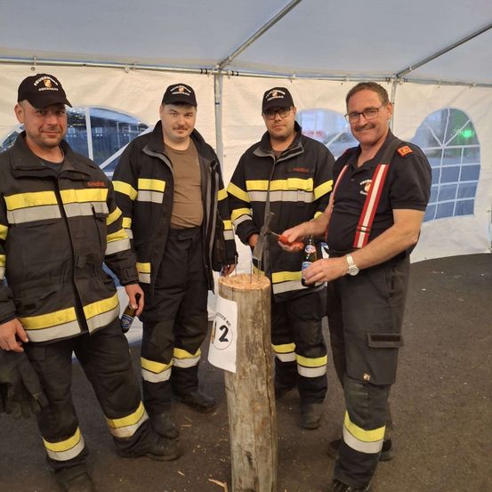 Vier Männer in Feuerwehruniformen stehen um einen Holzpfosten. Ein Mann hält eine Flasche und ein anderer einen Hammer. Sie lächeln.