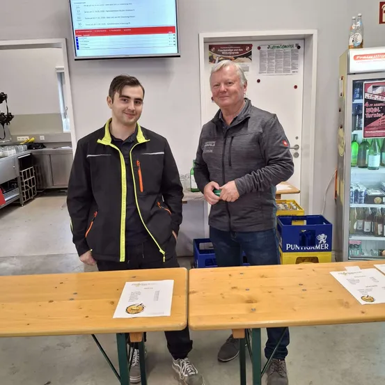 Zwei Männer stehen nebeneinander vor einem Tisch. Sie lächeln und scheinen für ein Foto zu posieren. Es liegen Papiere auf dem Tisch. Dahinter steht ein Kühlschrank mit Flaschen und einem Plakat. An der Wand ist ein Monitor montiert.
