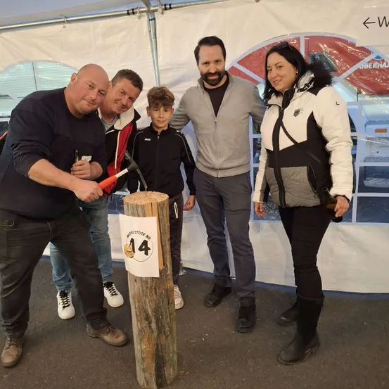 Fünf Personen posieren für ein Foto neben einem Holzpfosten mit der Nummer 14. Sie scheinen den Pfosten mit einem Vorschlaghammer zu schneiden.