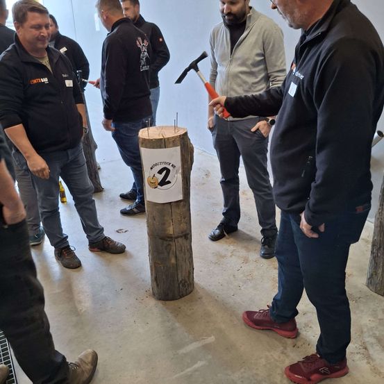 Eine Gruppe von Männern nimmt an einem Holzhackwettbewerb teil. Ein Mann hält einen Hammer und ist bereit, einen Holzstumpf mit der Nummer '2' zu treffen. Die anderen beobachten.