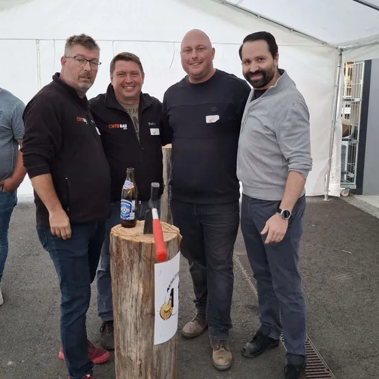 Vier Männer stehen um einen Baumstumpf mit einer Axt und einer Flasche. Sie scheinen für ein Foto zu posieren.