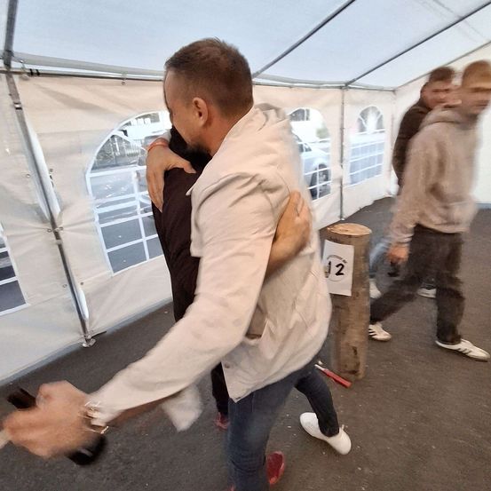 Ein Mann hält die Schulter und den Rücken eines anderen Mannes, während sie unter einem weißen Zelt gehen. Ein weiterer Mann geht hinter ihnen. Ein Holzstamm ist mit der Nummer 12 gekennzeichnet.