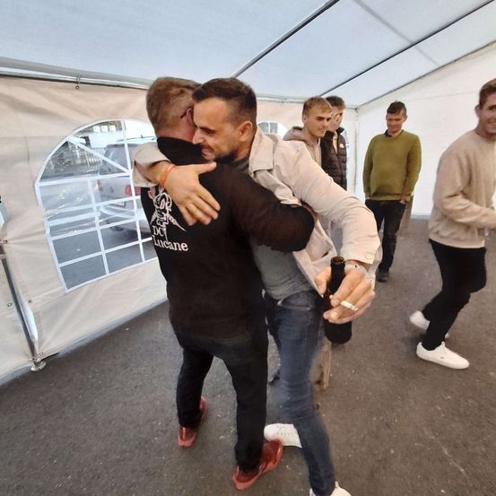 Zwei Männer umarmen sich in einem Zelt; einer hält ein Bier und trägt eine Uhr. Hinter ihnen gehen drei Männer, einer mit einem grünen Pullover. Vor dem Zelt ist ein Auto geparkt.