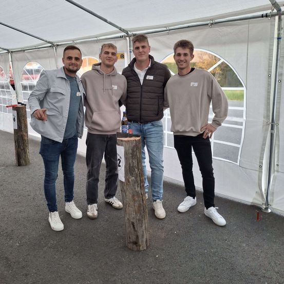 Vier Männer stehen lächelnd vor einem weißen Zelt. Ein Baumstumpf mit einer Flasche darauf steht zwischen ihnen. Sie tragen alle Freizeitkleidung. Das Zelt ist geöffnet, und ein Weg sowie ein Auto sind im Hintergrund zu sehen.