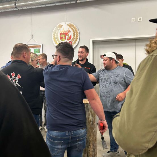 Eine Gruppe von Männern versammelt sich um einen Holzpfosten. Ein Mann hält ein Werkzeug und gestikuliert. Hinter ihnen eine Wand mit einem Logo und doppelten Türen.