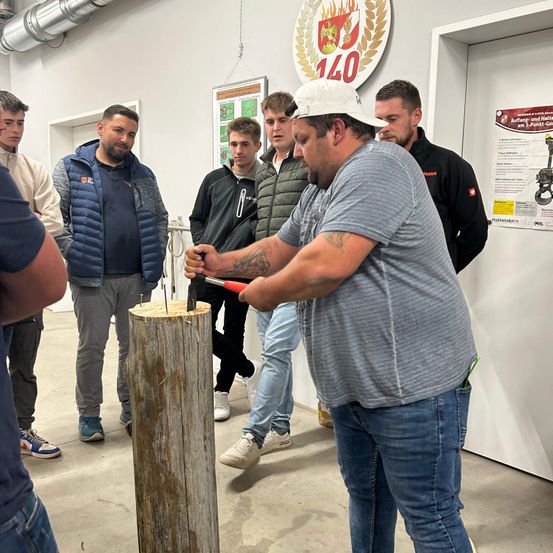 Ein Mann schlägt mit einem Hammer Nägel in einen Holzpfosten, während eine Gruppe von Männern zusieht.