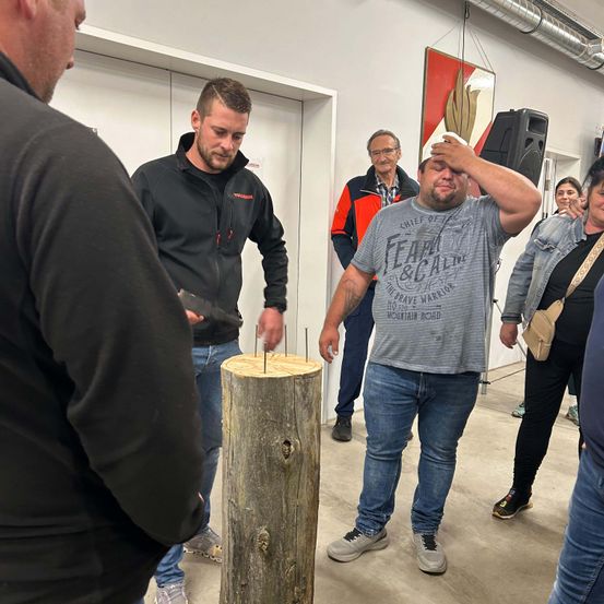 Ein Mann wirft einen Axt auf einen Holzstamm mit Nägeln, während andere zusehen, darunter ein Mann in einem grauen T-Shirt mit einem Bandana auf dem Kopf.