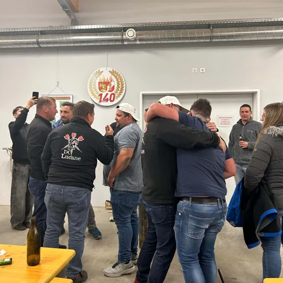 Eine Gruppe von Menschen in einem Raum, die feiern. Zwei umarmen sich, andere schauen zu. Es gibt einen Tisch mit einer Flasche und ein Schild mit der Nummer 140.