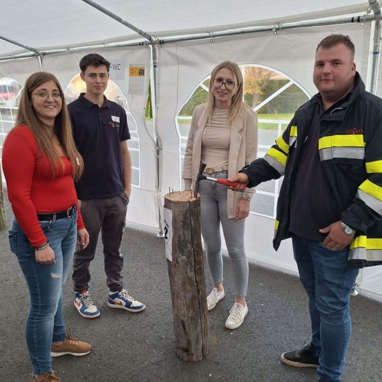 Vier Personen stehen unter einem weißen Zelt mit einem Baumstumpf in der Mitte. Der Mann auf der rechten Seite hält eine Säge. Die Frau in der Mitte trägt eine Brille. Ein Mann auf der linken Seite trägt Turnschuhe.