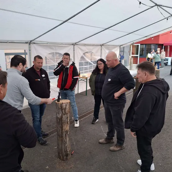 Eine Gruppe von Menschen steht in einem Zelt, einige von ihnen rauchen. Ein Mann hält einen Hammer und ist dabei, einen Baumstumpf zu treffen. Es stehen auch einige Leute außerhalb des Zeltes.