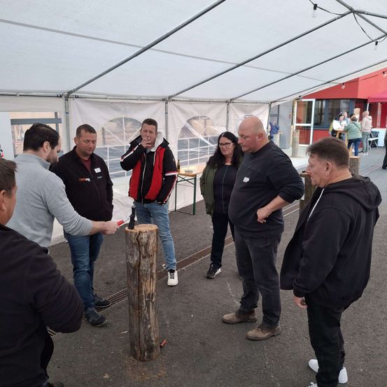 Eine Gruppe von Menschen steht in einem Zelt, einige von ihnen rauchen. Ein Mann hält einen Hammer und ist dabei, einen Baumstumpf zu treffen. Es stehen auch einige Leute außerhalb des Zeltes.