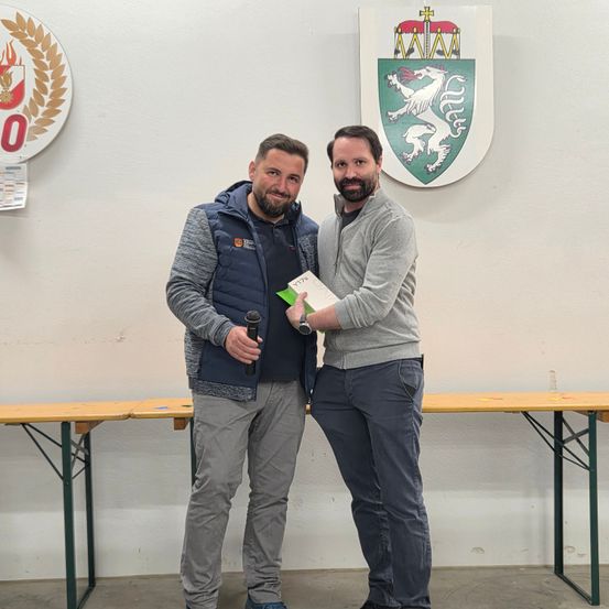 Zwei Männer stehen eng beieinander, einer hält ein Mikrofon und Papier, der andere hält Papier. Sie stehen vor einer Wand mit Emblemen und Tischen.