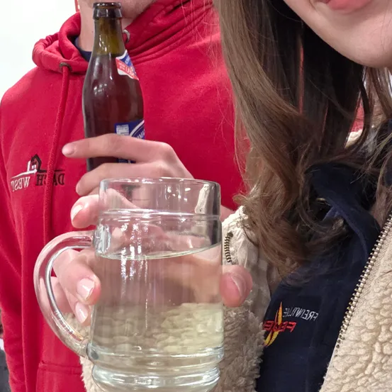 Eine Person in einer roten Kapuzenjacke hält ein Glas Wasser und eine Bierflasche. Eine andere Person in einer blauen Jacke lächelt.