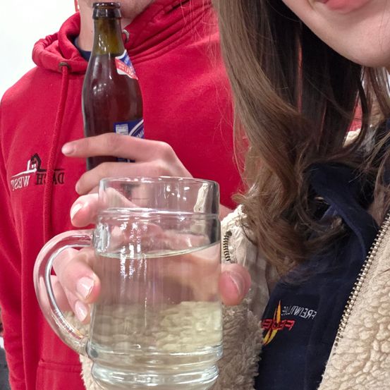 Eine Person in einer roten Kapuzenjacke hält ein Glas Wasser und eine Bierflasche. Eine andere Person in einer blauen Jacke lächelt.
