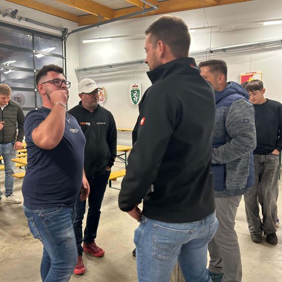 Mehrere Männer stehen in einem Raum mit Bänken und Mänteln. Ein Mann trägt eine Brille und berührt sein Kinn. Ein anderer Mann trägt einen Hut und Turnschuhe.