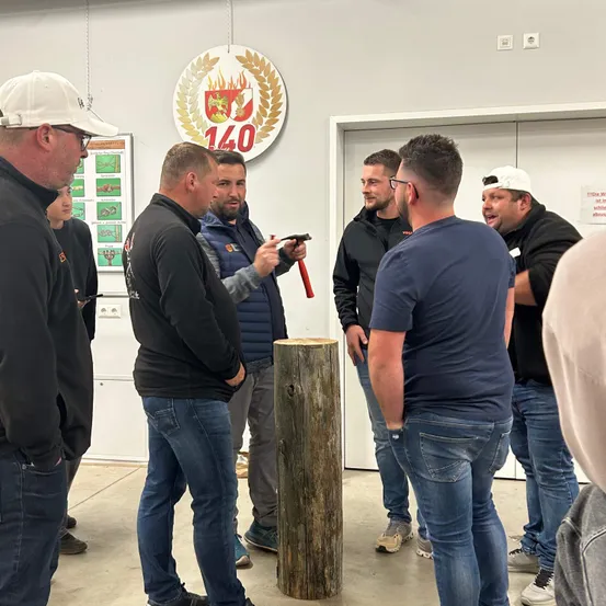 Fünf Männer stehen um einen Holzpfahl herum, einer hält einen Hammer, wahrscheinlich in einer Werkstatt. Ein rundes Logo mit der Nummer 140 ist über ihnen.
