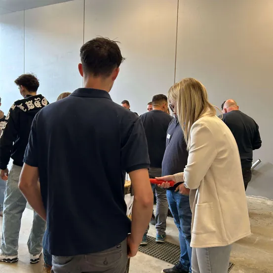 Eine Gruppe von Menschen in einem Flur, eine Frau hält ein rotes Objekt, möglicherweise ein Werkzeug, in ihren Händen.