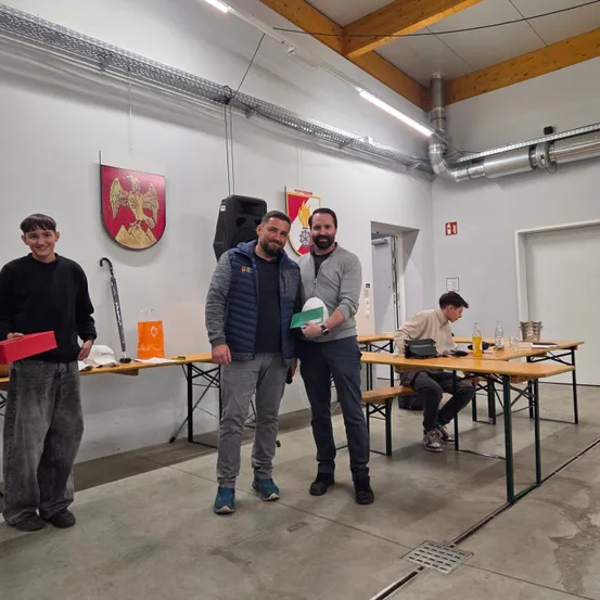 Drei Männer stehen in einem Raum mit Tischen und Bänken zusammen. Einer von ihnen hält eine rote Box. Ein Mann sitzt an einem der Tische.