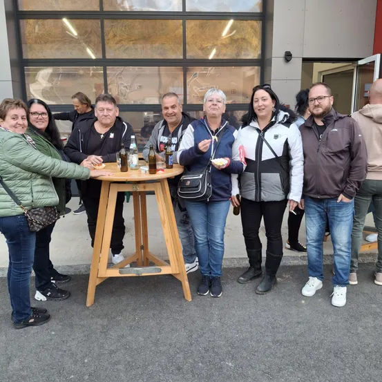 Eine Gruppe von Menschen, darunter Männer und Frauen, steht um einen runden Holztisch mit Getränken und Essen. Sie lächeln und scheinen für ein Foto zu posieren.