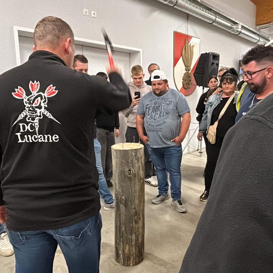 Eine Gruppe von Menschen steht um einen Holzpfahl herum. Ein Mann in einem DC Lucane Sweatshirt hält einen Hammer und ist dabei, zu schlagen. In der Nähe steht ein Mann in einem grauen Shirt und schaut zu.