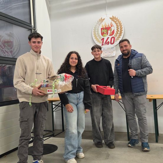 Vier Personen stehen in einem Raum mit einem Hintergrund, der ein Logo und die Nummer 140 zeigt. Die beiden Männer halten Geschenke, einer mit einem Mikrofon. Die Frau lächelt und hält ein Geschenk. Dahinter steht ein Tisch mit einem schwarzen Teppich.