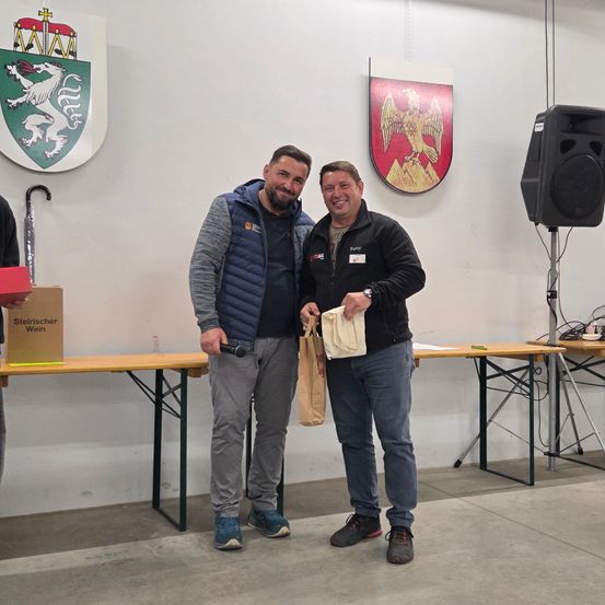 Zwei Männer stehen lächelnd neben einem Tisch, einer hält einen Papierbeutel. Hinter ihnen zeigt eine Wand zwei Wappen. Ein Lautsprecher steht auf einem Ständer rechts.