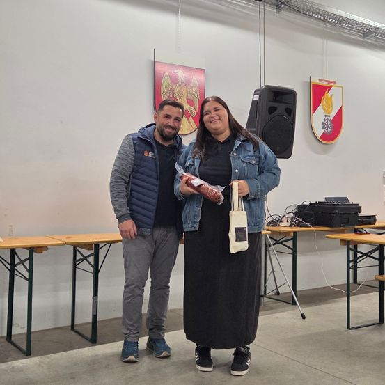 Ein Mann und eine Frau stehen nebeneinander in einem Raum mit Tischen und Bannern. Der Mann hält ein eingewickeltes Lebensmittel und die Frau einen weißen Beutel.