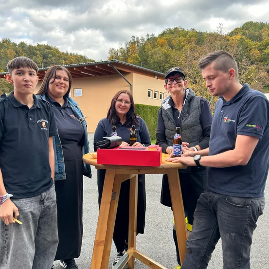 Fünf Personen stehen um einen holzfarbenen Tisch mit Bierflaschen und lächeln. Dahinter ist ein Gebäude mit einem geneigten Dach zu sehen, und es gibt Bäume und Büsche im Hintergrund.