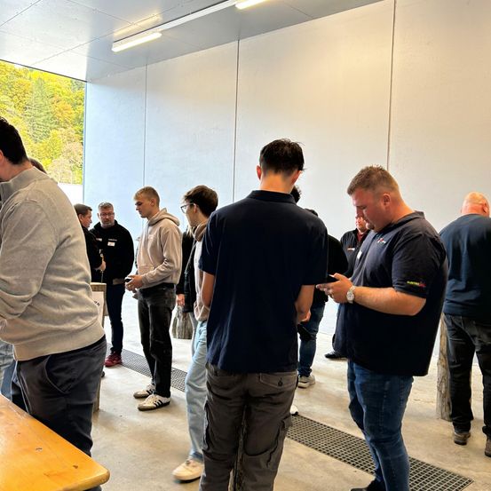 Eine Gruppe von Menschen ist drinnen versammelt, einige tragen Brillen, Turnschuhe und Uhren. Sie stehen in der Nähe eines Holztisches, mit einer Glaswand, die einen Blick auf Bäume bietet.