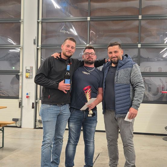 Drei Männer stehen zusammen in einem Raum, einer hält eine Trophäe und eine Flasche, während die anderen lässige Kleidung tragen. Sie scheinen eine Leistung zu feiern.