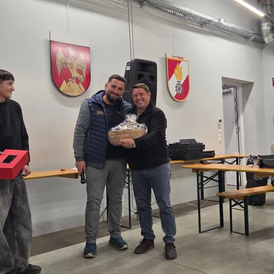Drei Männer stehen in einem Raum mit Tischen und einer Wand, die zwei Embleme zeigt. Zwei Männer halten einen Korb mit Essen und der dritte einen roten Kasten.