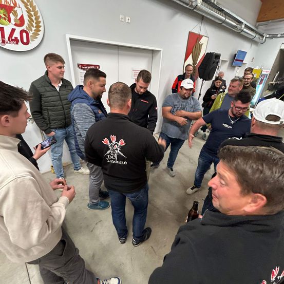Eine Gruppe von Männern steht in einem Raum, einige unterhalten sich miteinander. Ein Mann in einem schwarzen Shirt hält eine Flasche und schaut den Mann neben sich an. Ein Mann in einer weißen Kappe hält ein Handy. Hinter ihnen befindet sich eine Wand mit einem Logo.
