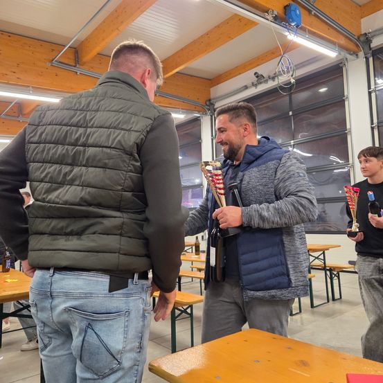 Zwei Männer stehen in einem Saal, einer präsentiert dem anderen einen Pokal. Der Mann mit dem Pokal lächelt und trägt eine Jacke. Hinter ihnen stehen Bänke und Tische, und ein Junge steht mit einem Pokal in der Hand.