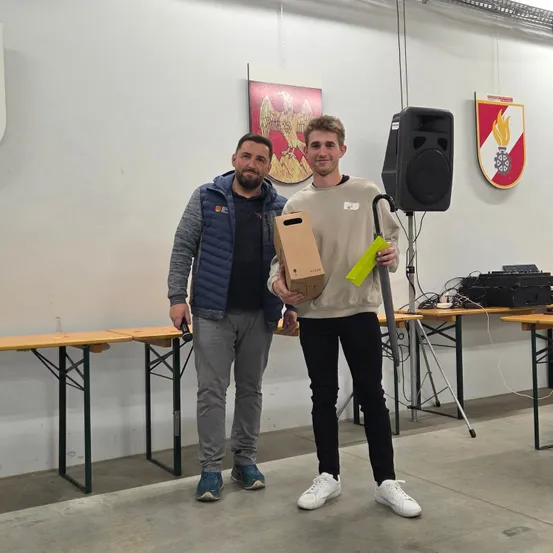 Zwei Männer stehen in einem Raum mit weißen Wänden. Einer hält eine Box und ein gelbes Band, der andere eine Flasche. Hinter ihnen steht ein Lautsprecher, ein Stativ und ein Banner. Tische und Stühle sind im Hintergrund aufgestellt.