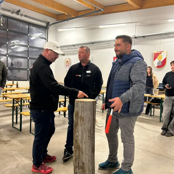 Drei Männer stehen um einen Holzpfosten in einem Raum mit Tischen und Bänken. Ein Mann hält einen Hammer und ein anderer zeigt auf den Pfosten. Ein Schild ist an der Wand hinter ihnen montiert.