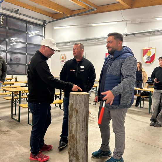 Drei Männer stehen um einen Holzpfosten in einem Raum mit Tischen und Bänken. Ein Mann hält einen Hammer und ein anderer zeigt auf den Pfosten. Ein Schild ist an der Wand hinter ihnen montiert.