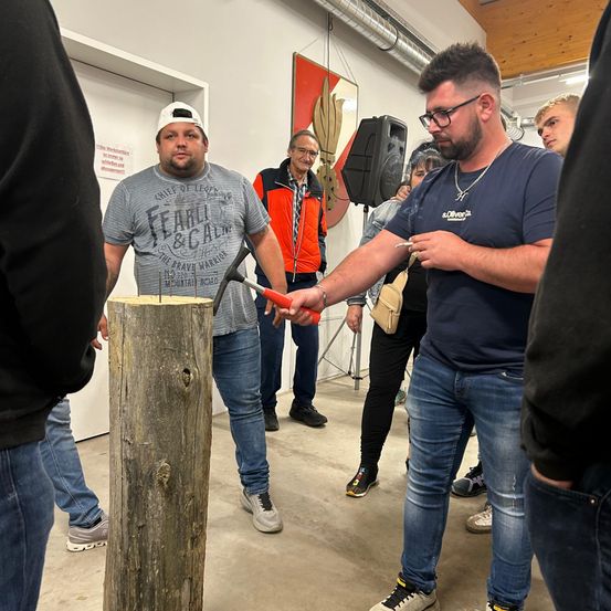 Ein Mann schlägt mit einem Hammer auf einen Baumstumpf, während ein anderer ihn festhält. Mehrere Leute beobachten, einer mit einer Zigarette. Im Hintergrund befinden sich ein Banner und ein Lautsprecher.