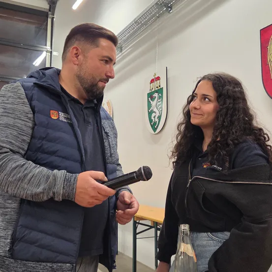 Ein Mann und eine Frau stehen in einem Raum und sehen sich an. Der Mann hält ein Mikrofon, was auf ein Interview oder ein Gespräch hindeutet. Hinter ihnen ist eine Wand mit Emblemen und in der Nähe steht eine Holzbank.