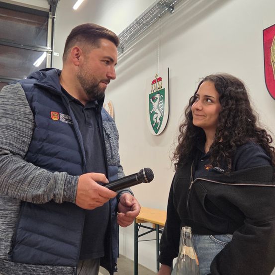 Ein Mann und eine Frau stehen in einem Raum und sehen sich an. Der Mann hält ein Mikrofon, was auf ein Interview oder ein Gespräch hindeutet. Hinter ihnen ist eine Wand mit Emblemen und in der Nähe steht eine Holzbank.