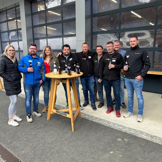 Eine Gruppe von Menschen steht draußen, lächelt und hält Flaschen auf einem Holztisch vor sich. Sie tragen Winterkleidung. Dahinter befindet sich ein Glasgebäude mit einem Logo.